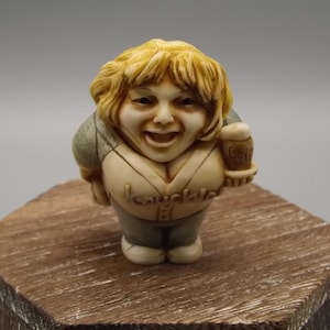 May include: A figurine of a smiling woman with blonde hair, wearing a beige top with the word "Knuckle" and gray sleeves. She holds a mug of beer. The figurine is on a wooden block.