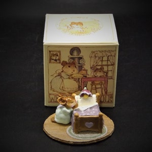 May include: A small ceramic figurine of a mouse in a nightgown kneeling beside a bed with a purple blanket and a heart-shaped pillow. The figurine is on a wooden base with a brown rug. The figurine is part of a collection called "The Mouse" and is packaged in a box with a picture of a mouse family.