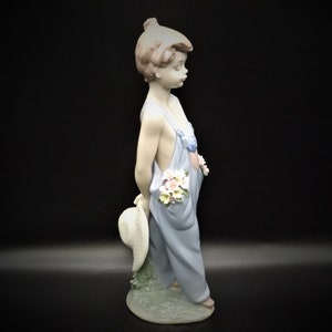 May include: A porcelain figurine of a young girl wearing a blue overall dress and holding a straw hat. She has flowers tucked into her overall pocket and is standing on a base with green grass.