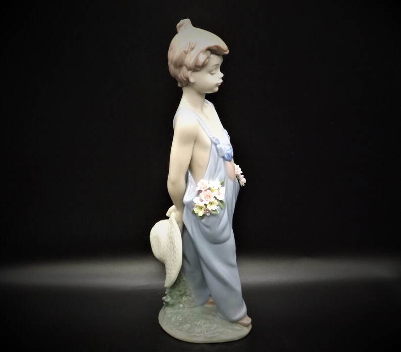 May include: A porcelain figurine of a young girl wearing a blue overall dress and holding a straw hat. She has flowers tucked into her overall pocket and is standing on a base with green grass.