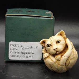 May include: A small, cream-colored figurine of a cat curled up in a ball. The figurine is made of ceramic and has a detailed, realistic appearance. The cat has large, blue eyes and a sweet expression. The figurine is sitting on a green box with the text "TJKIT01C 'Homer' Canadian Made in England for Harmony Kingdom".