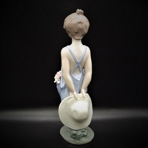 May include: A porcelain figurine of a girl wearing a blue dress and holding a straw hat. She has brown hair and is standing on a base with green leaves.