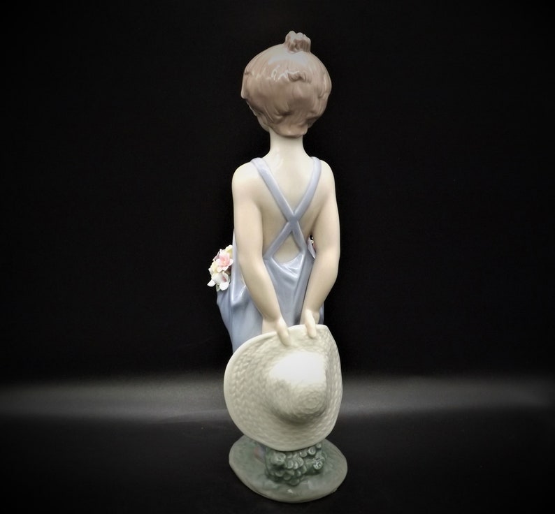 May include: A porcelain figurine of a girl wearing a blue dress and holding a straw hat. She has brown hair and is standing on a base with green leaves.