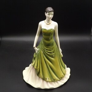 May include: A porcelain figurine of a woman in a strapless green gown with a white underskirt. The figurine has short dark hair, a gold necklace, and a bracelet. The dress has decorative gold accents.