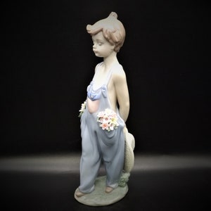 May include: A porcelain figurine of a young boy wearing blue overalls and holding a bouquet of flowers. He is standing on a base with green grass and flowers.