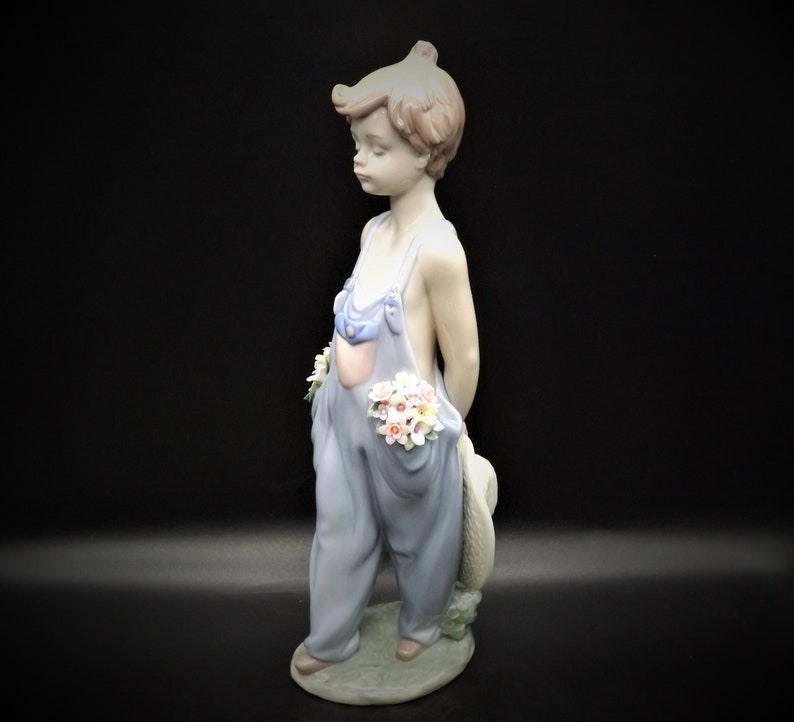 May include: A porcelain figurine of a young boy wearing blue overalls and holding a bouquet of flowers. He is standing on a base with green grass and flowers.