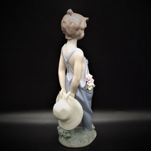 May include: A porcelain figurine of a young child wearing overalls and holding a straw hat. The child has pink flowers in their hair and is standing on a base with green leaves.