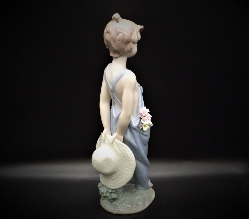May include: A porcelain figurine of a young child wearing overalls and holding a straw hat. The child has pink flowers in their hair and is standing on a base with green leaves.