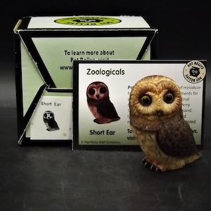 May include: A detailed figurine of a Short Ear owl, with brown and tan feathers, large eyes, and a small beak. The owl is displayed next to its box and a card with the text "Zoologicals" and "Short Ear".