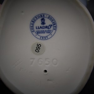 May include: White porcelain figurine base with a blue and white Lladro logo, the text "Collectors Society", "Hand Made in Spain", "DAISA 1996", and "1997". The base also has a white sticker with the number "095" and the number "7650" printed in black.