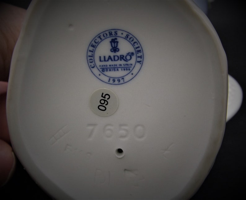 May include: White porcelain figurine base with a blue and white Lladro logo, the text "Collectors Society", "Hand Made in Spain", "DAISA 1996", and "1997". The base also has a white sticker with the number "095" and the number "7650" printed in black.