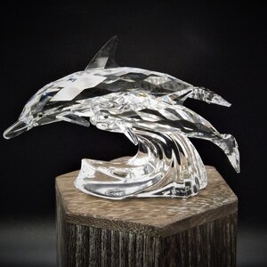 May include: A crystal sculpture of two dolphins leaping over a wave. The dolphins are made of clear crystal and have a faceted design. The wave is also made of clear crystal and has a flowing design.