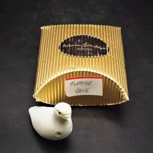 May include: A white ceramic turtle dove figurine with a small hole on its side, next to a gold corrugated box. The box has a label that reads "Adam Binder Editions" and a smaller label that says "Turtle Dove".