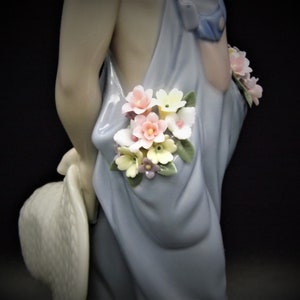 May include: A porcelain figurine of a girl wearing a blue dress with a floral design. The girl is holding a straw hat and has flowers in her hair.