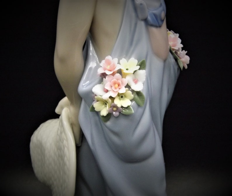 May include: A porcelain figurine of a girl wearing a blue dress with a floral design. The girl is holding a straw hat and has flowers in her hair.