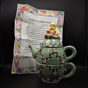 May include: A green ceramic teapot with a Tweety Bird figurine on top. The teapot is decorated with pink flowers and vines. The teapot is a three-piece set, including a teapot, a lid, and a cup. The certificate of authenticity reads: "This work of art is one of a limited signed edition by Southern California artist Laraine Eggleston. Each piece is unique because it is handpainted requiring countless hours of time and repeated firing. Attention to detail and the colors chosen to complement the design is the reason Laraine has collectors of her work in the United States and around the world."
