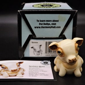 May include: A cream-coloured ceramic dog figurine named Spotsie, with brown accents, sits in front of its box. The box has text that reads "To learn more about Pot Bellys, visit www.HarmonyBall.com."