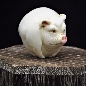May include: A white ceramic pig figurine with pink ears and snout. The pig is sitting on a wooden surface.