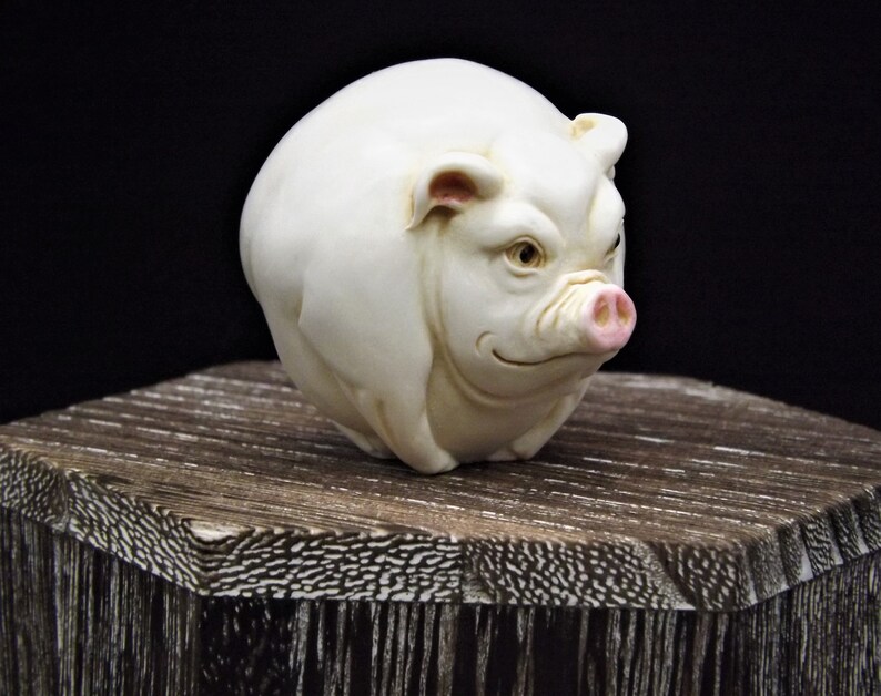 May include: A white ceramic pig figurine with pink ears and snout. The pig is sitting on a wooden surface.