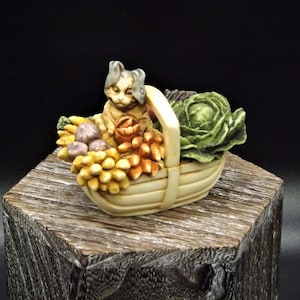May include: A ceramic figurine of a cat sitting in a basket filled with vegetables. The cat is gray and white with a green and brown basket. The vegetables include a cabbage, onions, and other vegetables.