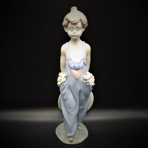 May include: A porcelain figurine of a young boy wearing blue overalls and holding a bouquet of flowers in each hand. He is standing on a base with green grass and a white hat.
