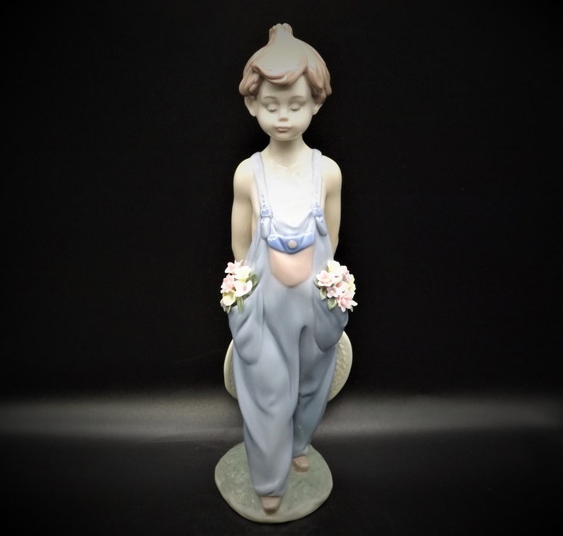 May include: A porcelain figurine of a young boy wearing blue overalls and holding a bouquet of flowers in each hand. He is standing on a base with green grass and a white hat.