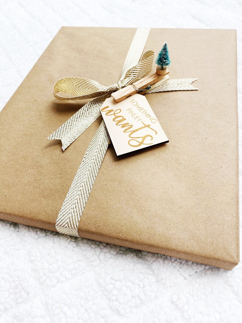 May include: A brown gift box wrapped with a gold ribbon and a wooden gift tag that says "Something Finley wants". A small green Christmas tree is attached to the tag with a wooden clothespin.