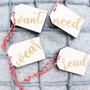 May include: Four white wooden gift tags with red and white twine. The tags have the words "want", "need", "wear", and "read" written in gold script.