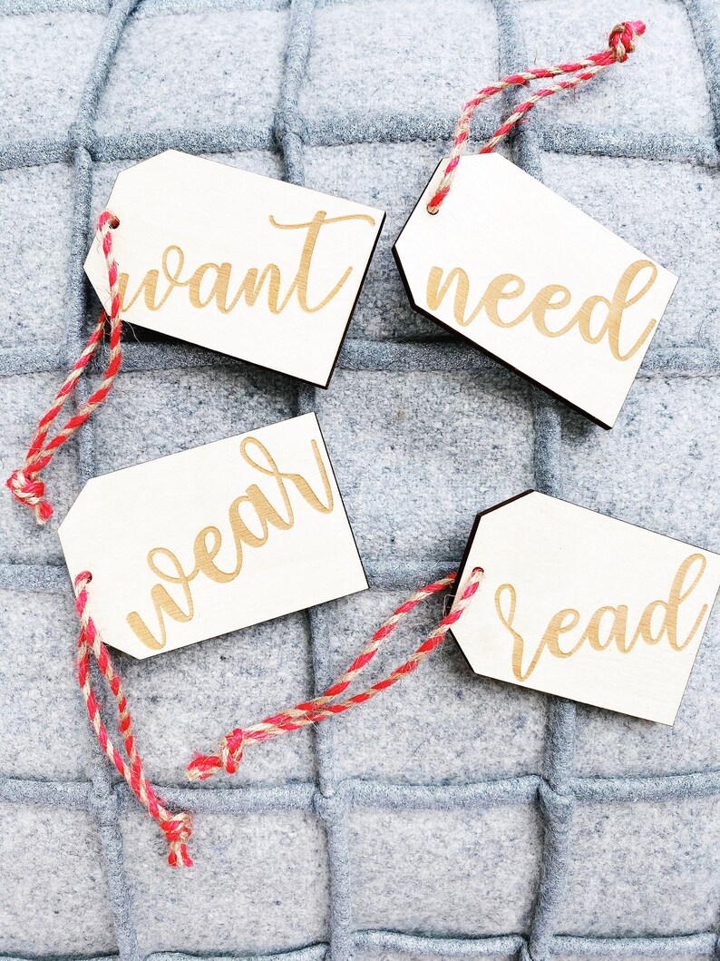 May include: Four white wooden gift tags with red and white twine. The tags have the words "want", "need", "wear", and "read" written in gold script.