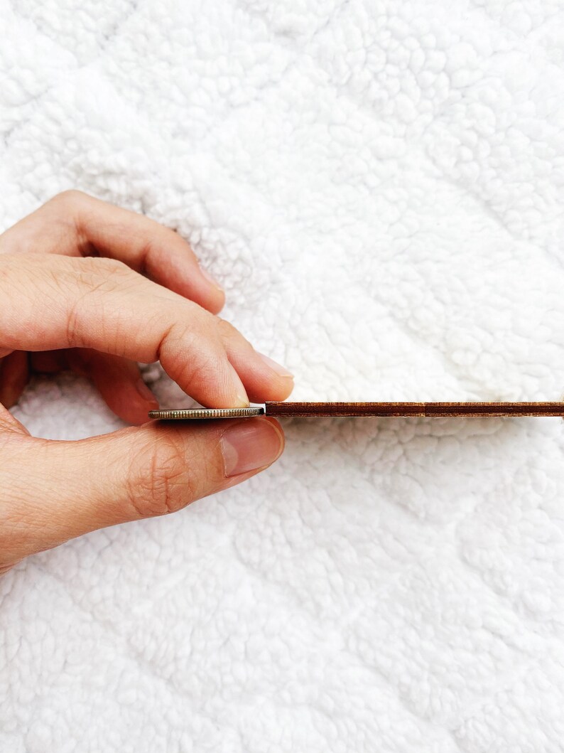 May include: A close-up of a wooden ruler being held by a hand. The ruler is brown and has a white edge. A coin is being used to hold the ruler in place.