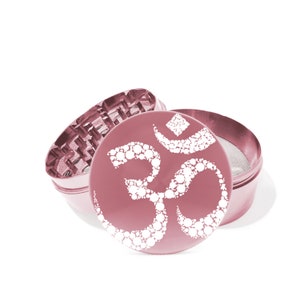 May include: A pink metal grinder with a white Om symbol design. The grinder has a three-piece design with a screen and a top lid.