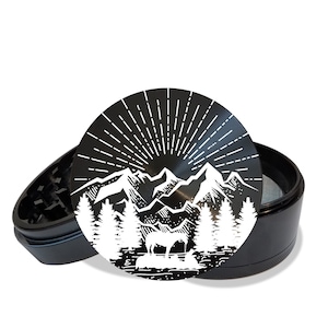 May include: Black metal grinder with a white mountain scene design. The design features a mountain range, trees, and a sunburst. The grinder is open and the top is off.