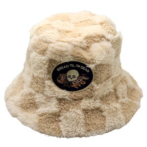 Op de afbeelding: Een beige en crème geblokte bucket hat gemaakt van zacht, pluizig materiaal. In het midden aan de voorzijde bevindt zich een ovaal embleem met een donkere achtergrond, voorzien van een geborduurde schedel met gebak en de tekst BREAD TIL I'M DEAD.