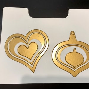 May include: A white sheet of paper with two gold metal die cuts. One die cut is a heart shape with three layers. The other die cut is a Christmas ornament shape with three layers.