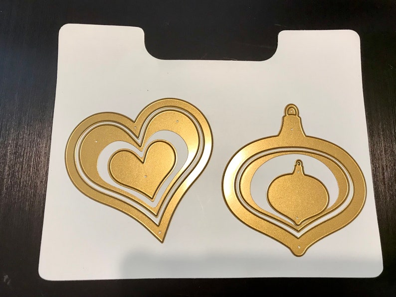 May include: A white sheet of paper with two gold metal die cuts. One die cut is a heart shape with three layers. The other die cut is a Christmas ornament shape with three layers.
