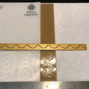 May include: A gold-colored metal die-cut border with a scalloped edge, positioned over a white embossed paper with a floral pattern. The image includes a Cricut Cuttlebug and Anna Griffin logo.