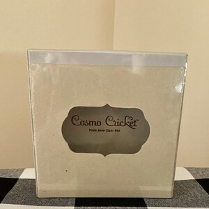 May include: A white cardboard box with a clear plastic window. The box has the text "Cosmo Cricket Plain Jane Cigar Box" printed on it.