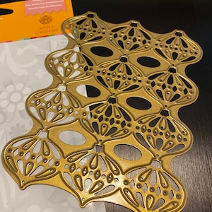 Puede incluir: Una troquelada de metal dorado con un dise&ntilde;o de ornamentos adornados superpuestos. La troquelada est&aacute; sobre un fondo blanco con un patr&oacute;n floral. La troquelada est&aacute; etiquetada como "Anna Griffin Ornament Dies".