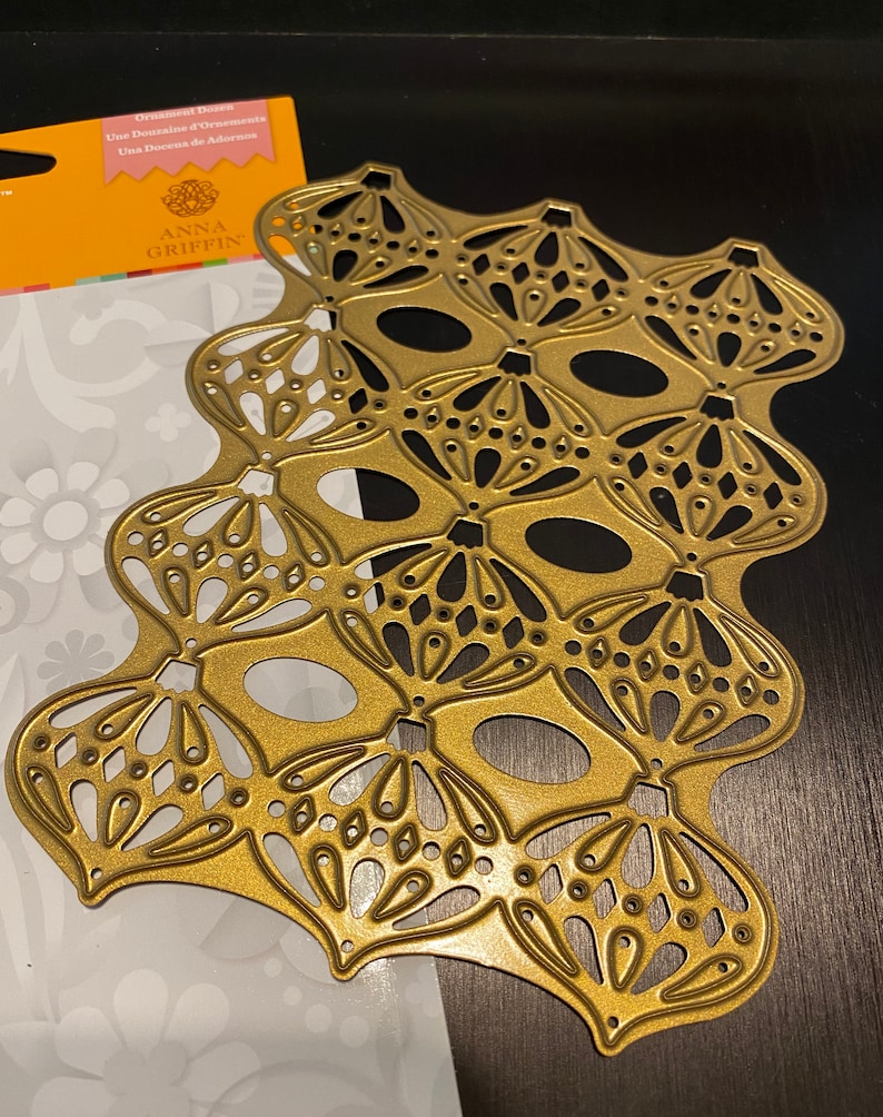 Puede incluir: Una troquelada de metal dorado con un dise&ntilde;o de ornamentos adornados superpuestos. La troquelada est&aacute; sobre un fondo blanco con un patr&oacute;n floral. La troquelada est&aacute; etiquetada como "Anna Griffin Ornament Dies".