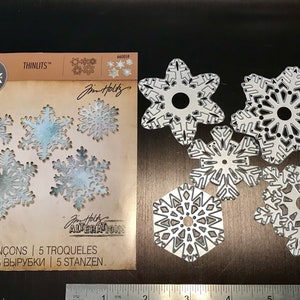 May include: A set of five snowflake-shaped metal dies for crafting. The dies are made by Sizzix and are part of the Jim Holtz Alterations collection. The dies are silver in color and have intricate designs.