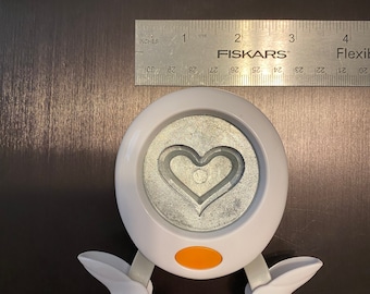 May include: White heart-shaped paper punch with orange handle. The punch is designed to create a double heart shape.