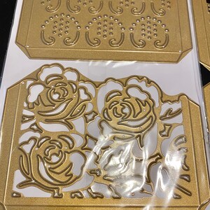 Puede incluir: Dos plantillas de metal dorado troqueladas, una con un dise&ntilde;o floral y la otra con un patr&oacute;n repetitivo de puntos y espirales.