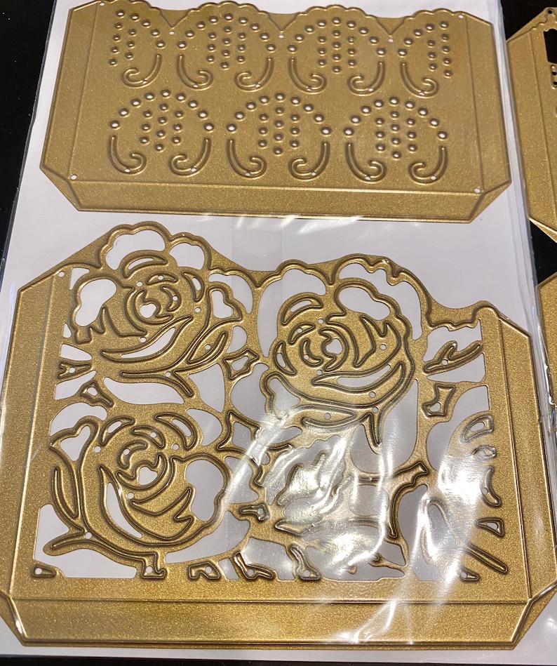 Puede incluir: Dos plantillas de metal dorado troqueladas, una con un dise&ntilde;o floral y la otra con un patr&oacute;n repetitivo de puntos y espirales.