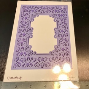 May include: Purple die-cutting template with a decorative frame and intricate floral design. The template is made by Cutztlebug.