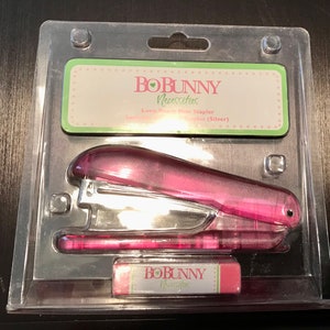 May include: A pink plastic stapler with a silver staple remover and a pink eraser with the text "BoBunny Necessities" on it. The stapler is packaged in a clear plastic clamshell.