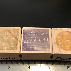 May include: Three wooden rubber stamps with different designs. The first stamp has a green and white design with Chinese characters. The second stamp has a purple and white design with musical notes. The third stamp has a yellow and red design with a clock face. All three stamps have the word "Musto" written on the bottom.