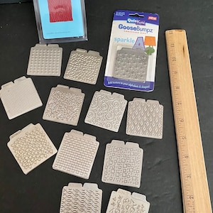 May include: A set of 10 silver metal embossing dies for crafting. The dies feature various patterns, including stars, swirls, hearts, and geometric shapes. The dies are labeled with the brand name "GooseBumpz" and the phrase "sparkle".