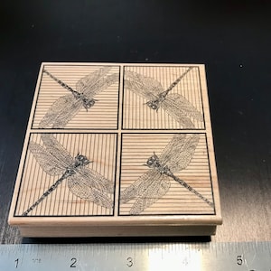 May include: A wooden stamp with four squares, each featuring a black and white illustration of a dragonfly with detailed wings. The dragonfly is set against a background of thin, horizontal lines.