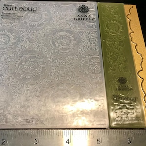 May include: A clear plastic embossing plate with a floral pattern, a green plastic embossing plate with a floral pattern, and a gold plastic border. The plates are labeled "Cricut cuttlebug Blossom Topaz Glissement de fleurs  Mecia de Flores" and "Anna Griffin".