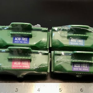 May include: Four green plastic containers with labels that say "Acid Free" and the color of the page inside. The labels also say "Ancient Page" and the color of the page. The containers are lined up in a row on a black surface with a ruler underneath.